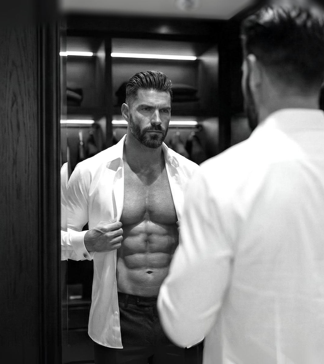 Man with a beard and muscular build wearing a white shirt, looking at himself in a mirror.